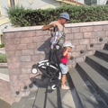 A father holds up a stroller while his toddler walks up stairs with it still attached to her