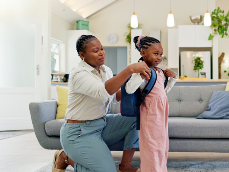 A happy mother gets her toddler ready for school by helping her with her backpack