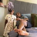 A little girl stands next to her father while he lies on his bed