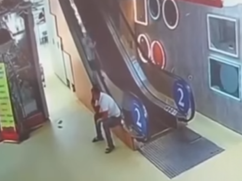 A man is on the phone while his child goes up the side of an escalator
