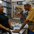 A man uses a hot sauce bottle as a mic and sings for a cashier at the checkout