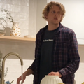 A man washes dishes and his eyes are glued to the TV