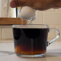 A person cracks a whole egg into a cup of black coffee