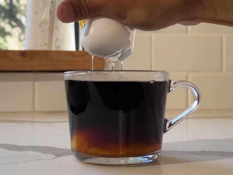 A person cracks a whole egg into a cup of black coffee