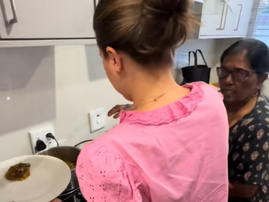 A woman dishing out food while an older Indian woman shows her what she has cooked