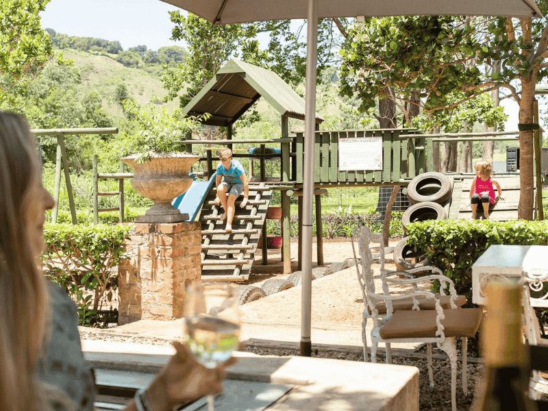 A woman holds a glass of wine while looking at her kids playing at play area in restaurant