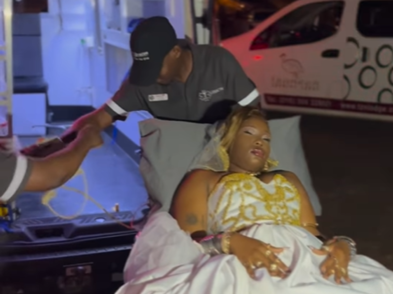A young woman wearing a ballgown lies on a stretcher outside an ambulance van