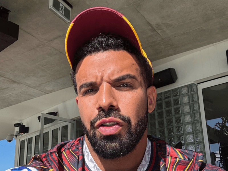 Drake wears a red and yellow baseball cap while taking a selfie